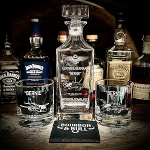 May include: A clear glass decanter with a personalized inscription for "CDR Mike Bernar "Bernie" - EA-18G Growler - Mission Complete". Two matching glasses with the same inscription are also shown. The decanter and glasses are sitting on a black coaster with the words "Bourbon & Bull" in white.