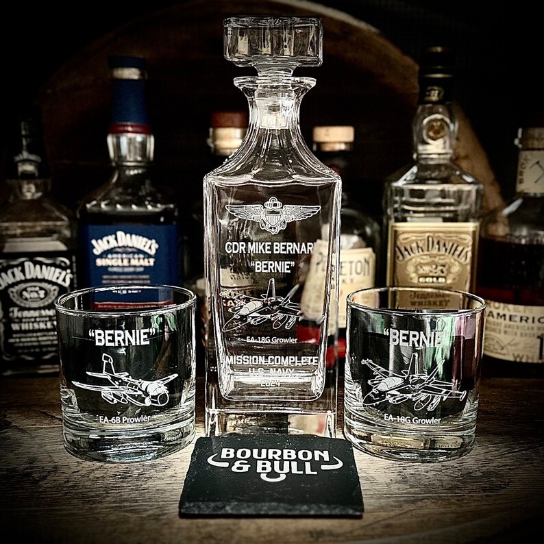 May include: A clear glass decanter with a personalized inscription for "CDR Mike Bernar "Bernie" - EA-18G Growler - Mission Complete". Two matching glasses with the same inscription are also shown. The decanter and glasses are sitting on a black coaster with the words "Bourbon & Bull" in white.