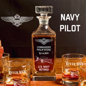 May include: A clear glass decanter with a square base and a stopper. The decanter is engraved with the words "Commander Philip Stone", "River Man", "U.S. Navy Retired", and a Navy pilot's wings. The decanter is surrounded by three clear glass tumblers, each engraved with the words "River Man" and a Navy pilot's wings.