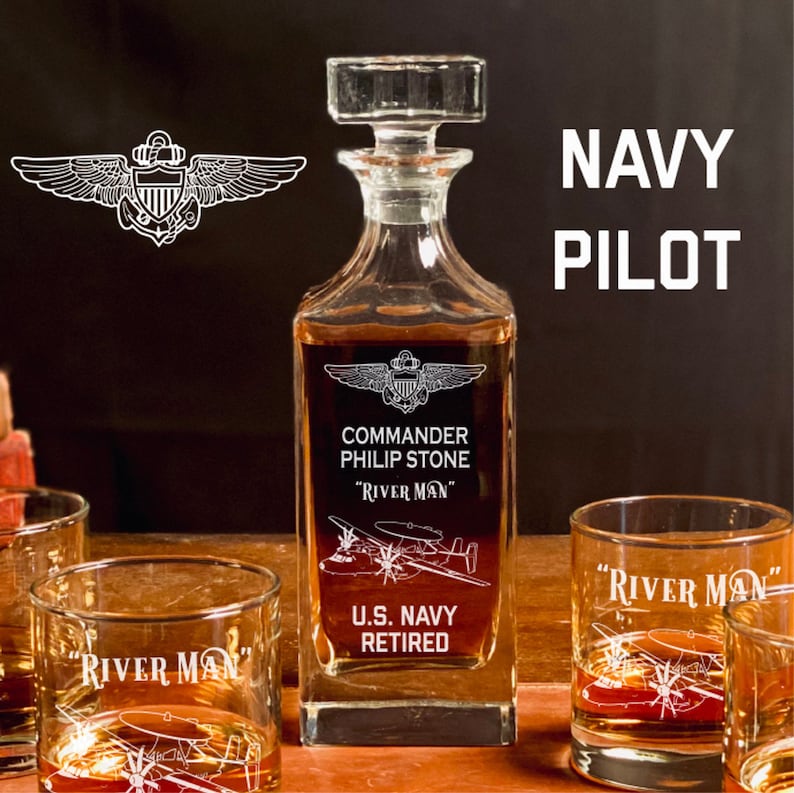 May include: A clear glass decanter with a square base and a stopper. The decanter is engraved with the words "Commander Philip Stone", "River Man", "U.S. Navy Retired", and a Navy pilot's wings. The decanter is surrounded by three clear glass tumblers, each engraved with the words "River Man" and a Navy pilot's wings.