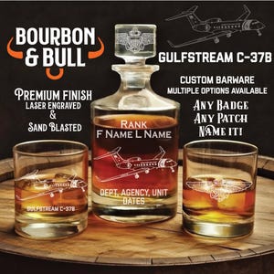 May include: A custom barware set featuring a decanter and two glasses, laser engraved and sandblasted. The decanter has a stopper with a crest design. The glasses and decanter are etched with a Gulfstream C-37B airplane. Text includes "Bourbon & Bull" and "Custom Barware".