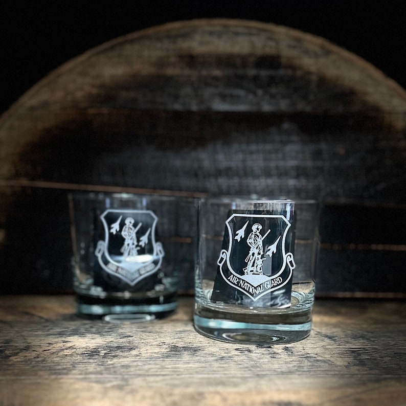 Puede incluir: Dos vasos de vidrio transparente con un dise&ntilde;o grabado en blanco del logotipo de la Guardia Nacional A&eacute;rea. El logotipo presenta a un soldado sosteniendo un rifle y dos aviones a reacci&oacute;n en el fondo.