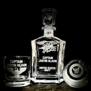 May include: A clear glass decanter with a matching tumbler and a glass lid. The decanter and tumbler are etched with the words "CAPTAIN JUSTIN HLAVIN" and "UNITED STATES NAVY" along with a military emblem. The lid also has an emblem.