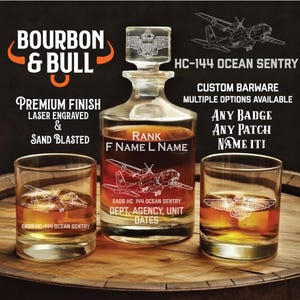 May include: A clear glass decanter and two matching glasses with etched designs. The decanter has a stopper and is labeled "Bourbon & Bull." The glasses and decanter feature an airplane design and the text "HC-144 OCEAN SENTRY."
