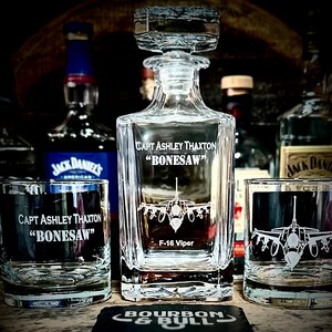 May include: A personalized crystal decanter and two matching glasses with an etched design of an F-16 Viper fighter jet. The decanter and glasses are engraved with the text "CAPT ASHLEY THAXTON "BONESAW" F-16 Viper".