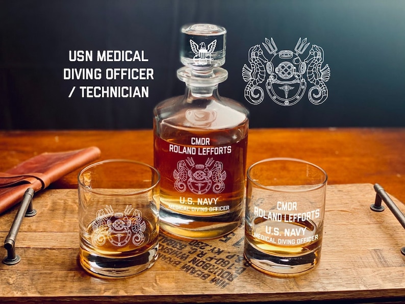 May include: A set of two clear glass tumblers and a decanter with a stopper. The glasses and decanter are engraved with the text "CMDR ROLAND LEFFORTS U.S. NAVY MEDICAL DIVING OFFICER" and a nautical emblem featuring a diving helmet, seahorses, and tridents.