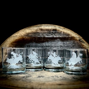 May include: Set of four clear glass whiskey tumblers with an etched design of a cowboy riding a bull. The glasses are arranged in a row on a wooden surface.