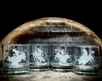May include: Set of four clear glass whiskey tumblers with an etched design of a cowboy riding a bull. The glasses are arranged in a row on a wooden surface.