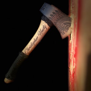 Op de afbeelding: Bijl met een houten handvat en een donkere metalen kop met een gegraveerd Indiaanse hoofdtooi-ontwerp met een schedel. De handgreep is gegraveerd met "Christopher Hasty". De bijl is gedeeltelijk ingebed in een rood-wit oppervlak.