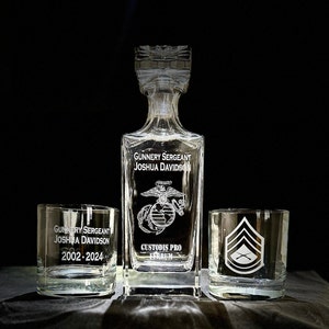 May include: A set of three clear glass decanters with etched designs. The decanter in the center has the text "Gunnery Sergeant Joshua Davidson" and a military emblem. The decanters on either side have the text "Gunnery Sergeant Joshua Davidson 2002-2024" and a military rank insignia.
