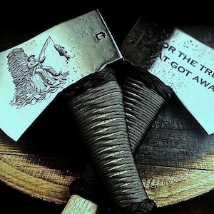 May include: Two axes with black wrapped handles rest on a wooden surface. The axe heads have etched designs. One shows a man fishing, the other reads "FOR THE TREE THAT GOT AWAY."