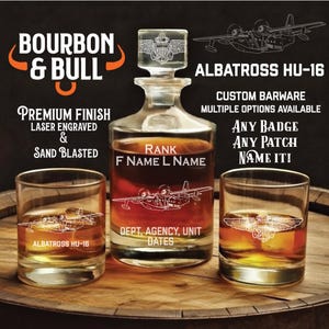May include: A custom barware set featuring a decanter and two glasses, laser engraved and sandblasted. The set includes the text "BOURBON & BULL" and "ALBATROSS HU-16". The decanter and glasses are clear, and the liquid inside is amber.