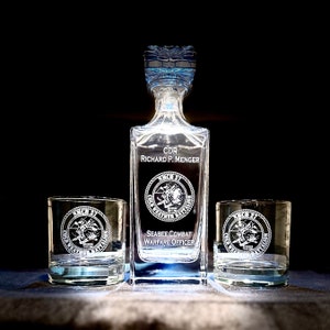 May include: A set of three clear glass decanters with engraved text and a military insignia. The decanters are on a dark surface. The text reads "CDR Richard P. Menger" and "Seabee Combat Warfare Officer".