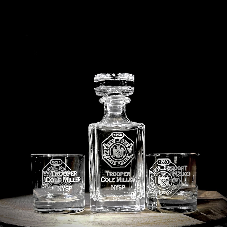 May include: A set of three clear glass decanters with etched designs. The decanters feature the New York State Police logo and the text "Trooper Cole Miller NYSP".