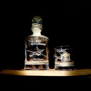May include: A clear glass decanter and a matching glass tumbler with an engraved design of a military aircraft and the text "MSGT ZACH LEMASTER" and "MQ-9 Reaper" on the decanter and "CHAZZ" and "MQ-9 Reaper" on the tumbler.