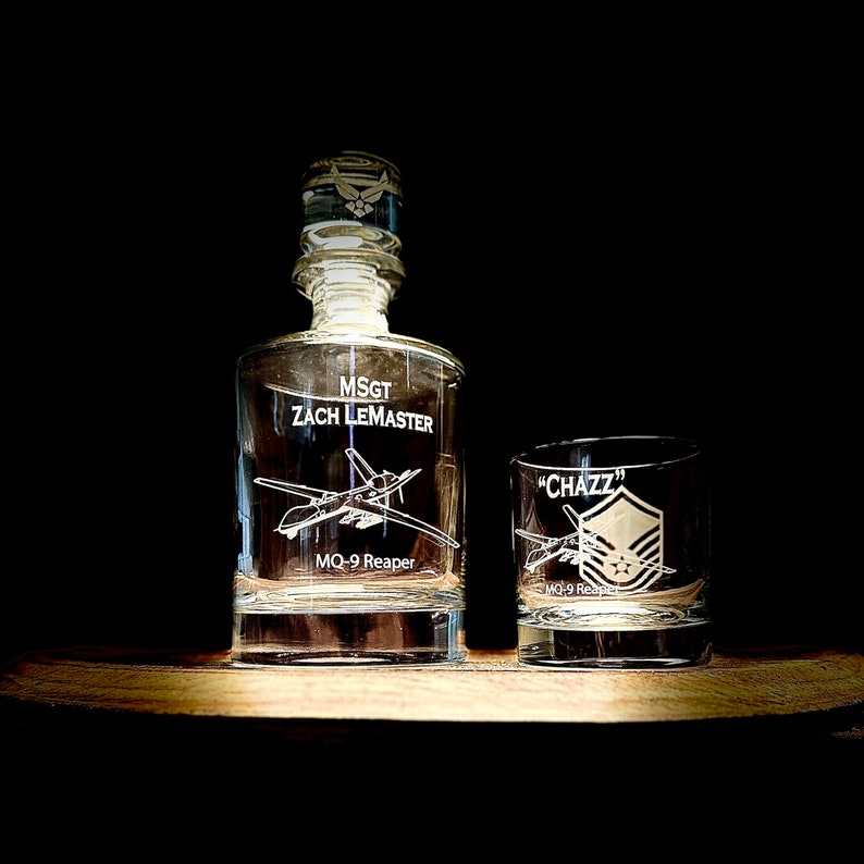 May include: A clear glass decanter and a matching glass tumbler with an engraved design of a military aircraft and the text "MSGT ZACH LEMASTER" and "MQ-9 Reaper" on the decanter and "CHAZZ" and "MQ-9 Reaper" on the tumbler.