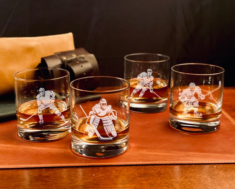 Puede incluir: Cuatro vasos transparentes de vidrio con dise&ntilde;os de jugadores de hockey grabados. Los vasos est&aacute;n llenos de un l&iacute;quido marr&oacute;n y est&aacute;n sobre una superficie de cuero marr&oacute;n.