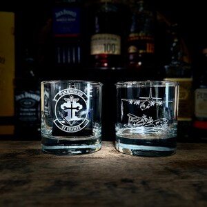 May include: Two clear glass tumblers with etched designs. One tumbler features a military insignia with the text "Para Bellum 27 SOAMXS". The other tumbler features an etched image of a military aircraft.