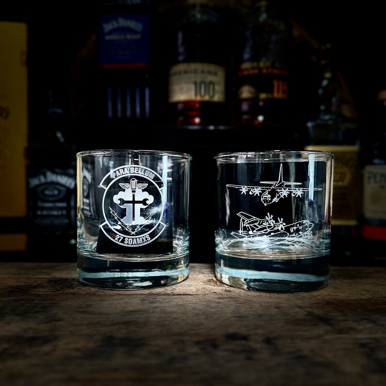 May include: Two clear glass tumblers with etched designs. One tumbler features a military insignia with the text "Para Bellum 27 SOAMXS". The other tumbler features an etched image of a military aircraft.