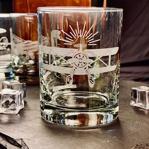 May include: A clear glass tumbler featuring a white etched design of a biplane with a propeller and a sunburst pattern above it. The glass has a thick base and is cylindrical in shape, showcasing a detailed front-facing view of the aircraft.
