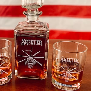May include: A set of three clear glass decanters with a helicopter design and the word "Skeeter" etched on them. The decanters are filled with a dark liquid and are sitting on a wooden surface. The decanter in the center has the text "MH-53J Pave Low" etched on it.