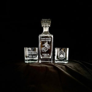 May include: A set of three clear glass decanters with etched designs. The largest decanter is a liquor decanter with the text "Gunnery Sergeant Joshua Davidson Custodis Pro Ferrum" and a USMC emblem. The two smaller decanters are whiskey glasses with the text "Gunnery Sergeant Joshua Davidson 2002-2024" and a USMC rank insignia.