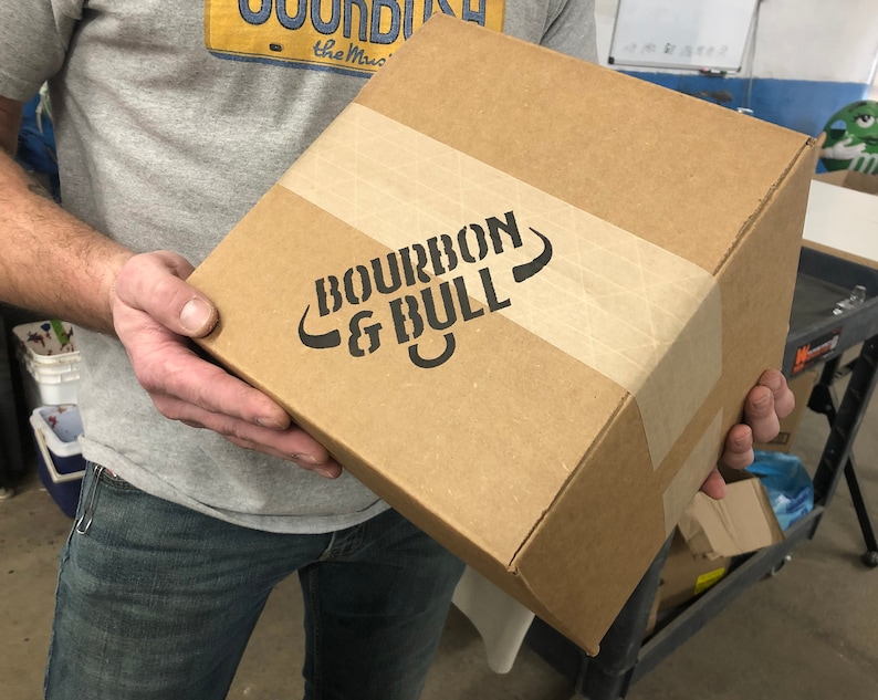Puede incluir: Una caja de cart&oacute;n marr&oacute;n con las palabras "BOURBON & BULL" impresas en negro en el lateral. La caja est&aacute; cerrada con cinta adhesiva y tiene una etiqueta de papel marr&oacute;n.