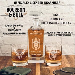 May include: A clear glass decanter and two matching glasses filled with amber liquid. The decanter and glasses are laser engraved with a military insignia and text. The text includes "BOURBON & BULL", "USAF COMMAND CHIEF MASTER SERGEANT", and "U.S. AIR FORCE".
