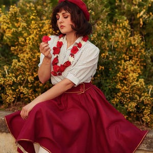Pu&ograve; includere: Una donna indossa un berretto rosso, una blusa bianca a pois, una collana di fiori rossi e una gonna bordeaux con un bordo dorato. Tiene un fiore rosso in mano ed &egrave; seduta davanti a un cespuglio di fiori gialli.