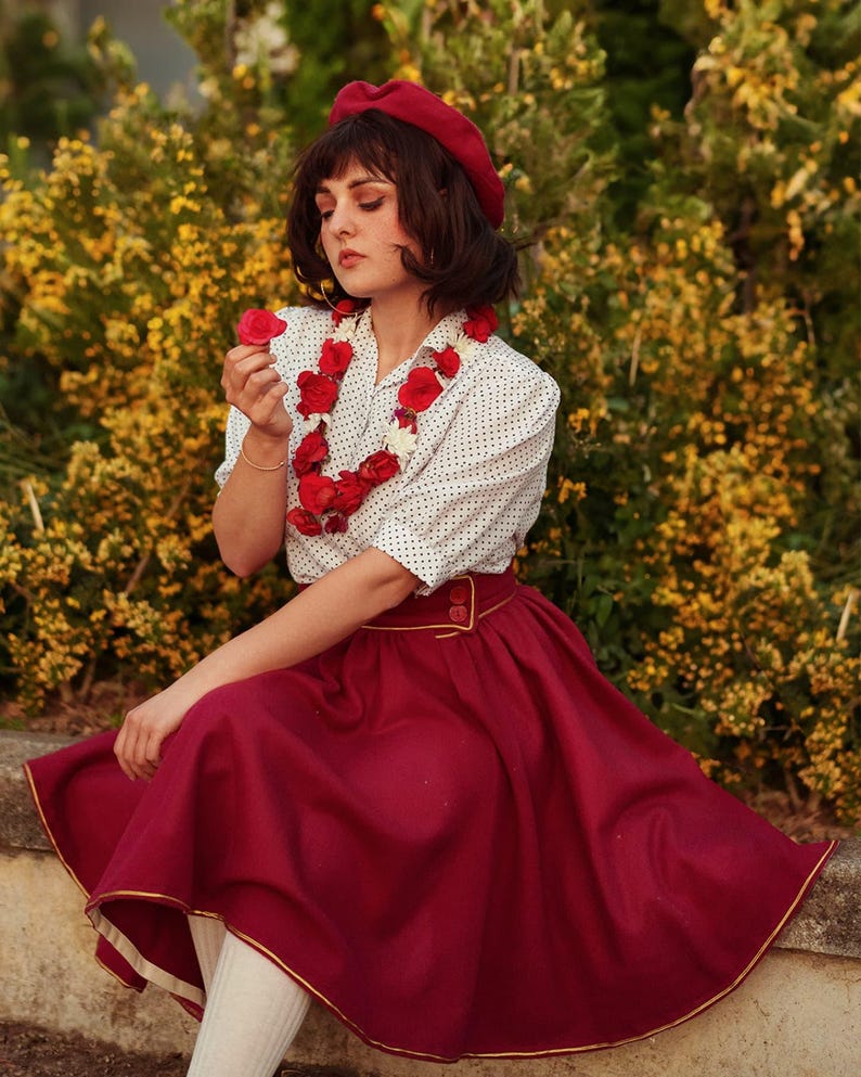 Pu&ograve; includere: Una donna indossa un berretto rosso, una blusa bianca a pois, una collana di fiori rossi e una gonna bordeaux con un bordo dorato. Tiene un fiore rosso in mano ed &egrave; seduta davanti a un cespuglio di fiori gialli.