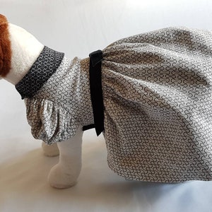 May include: A white and black patterned dog dress with a black bow. The dress has a ruffled collar and a full skirt.
