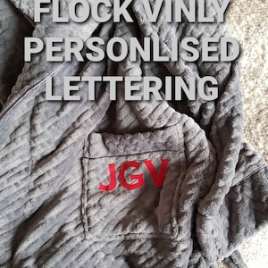 Op de afbeelding: Grijze badjas met capuchon met een zak op de voorkant. De zak heeft rode flock vinyl letters met de tekst "JGV".