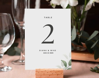 May include: White table number card with the text "TABLE 2" in black. Below the number is the text "DIANA & MIKE AUG 22 2025" in black. The card has a watercolor design of green leaves in the bottom right corner. The card is sitting on a wooden stand.