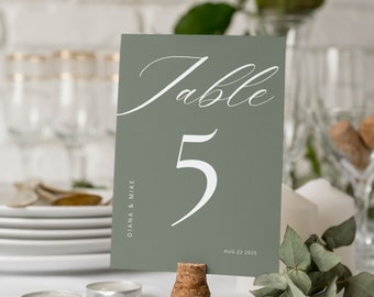 May include: Sage green table number card with white calligraphy that says "Table 5" and the date "Aug 22 2025". The card is on a cork stand and there are small candles and greenery on the table.