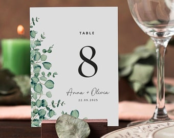May include: A white table number card with a green eucalyptus leaf design. The card has the text "TABLE 8" and "Anna + Olivia 22.09.2025".