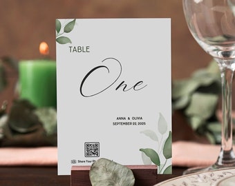 May include: A white table number card with green foliage and the text "TABLE One" and "ANNA & OLIVIA SEPTEMBER 22, 2025". A QR code is included with the text "Share Your Photos". The card is on a wooden stand with a green leaf.