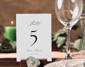 May include: White table number card with black calligraphy that reads "Table 5" and the text "Anna + Olivia 22.09.2025". The card is propped up on a small wooden stand with a green leaf.