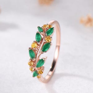 May include: A rose gold ring featuring a branch-like design adorned with marquise-cut green gemstones and small round yellow gemstones arranged in a leaf pattern along the top half of the thin, polished metal band.