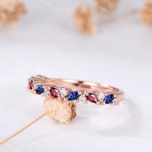 May include: Rose gold ring with alternating red, blue, and white gemstones. The gemstones are marquise-cut and set in a row along the band. The ring is displayed on a white surface.