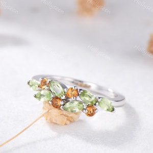 May include: A silver ring featuring a cluster of green and orange gemstones. The ring's design incorporates marquise-cut green stones and round orange stones, creating a nature-inspired aesthetic. The ring is set against a soft, white background.
