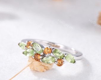 May include: A silver ring featuring a cluster of green and orange gemstones. The ring's design incorporates marquise-cut green stones and round orange stones, creating a nature-inspired aesthetic. The ring is set against a soft, white background.