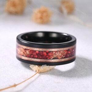 May include: A black ring with a red and gold inlay. The ring has a wide band with a black exterior and a copper-coloured band bordering the inlay. The inlay features red and gold-coloured fragments.