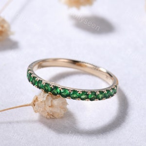 Puede incluir: Un delicado anillo de banda de oro con una fila de pequeñas piedras preciosas verdes. El anillo está sobre un fondo blanco.