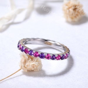 May include: A delicate eternity band ring with a row of alternating pink and purple gemstones. The ring is made of white metal and has a polished finish.