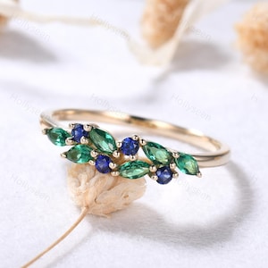 May include: A gold ring featuring a curved arrangement of marquise-cut green stones and round-cut blue stones. The band is thin and polished, with the gemstones set in a delicate, leaf-like pattern across the top of the ring's circular frame.