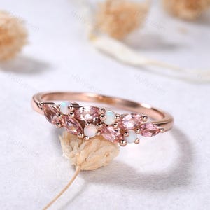 May include: Rose gold ring with a row of pink marquise-cut gemstones and white opal stones. The ring is set on a delicate band, with the gemstones arranged in a floral or leaf-like pattern. The ring is displayed on a white surface.