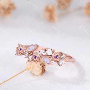 May include: A rose gold ring featuring a floral design with small purple stones at the center of each flower, clear round accent stones, and two marquise-shaped iridescent opal-like stones arranged along the band in a nature-inspired pattern.