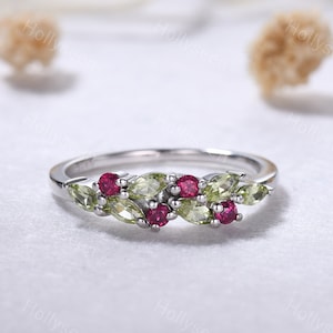 May include: A silver ring with a delicate design featuring a row of alternating green and red gemstones. The ring is set with small, round red gemstones and pear-shaped green gemstones.