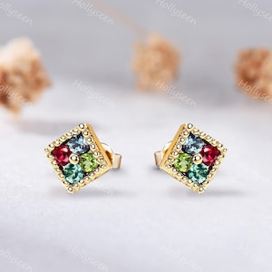 May include: A pair of gold stud earrings, each featuring a square frame with a beaded edge. Inside each square are four round-cut gemstones: one red, one light green, one blue-green, and one grayish-blue, arranged in a cluster setting.