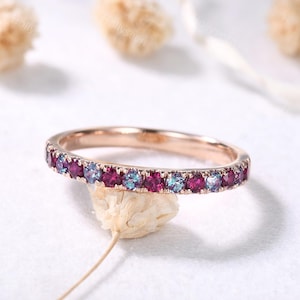 May include: Rose gold ring with alternating ruby and alexandrite gemstones. The ring features a thin band and a continuous row of vibrant, colorful stones. The gemstones are a mix of deep red and teal blue, set in a polished metal band.